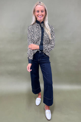 Woman wearing a patterned jacket, black top, dark jeans, and white shoes on a plain background