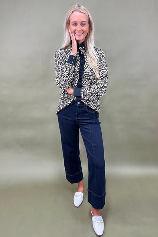 Woman wearing a patterned blazer, dark jeans, and white shoes on a plain background