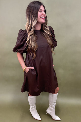 Woman wearing a brown dress with puffed sleeves and white boots against a green background
