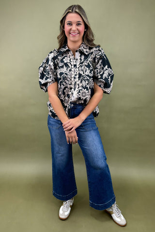 Woman wearing a black and white floral blouse with blue jeans against a green background