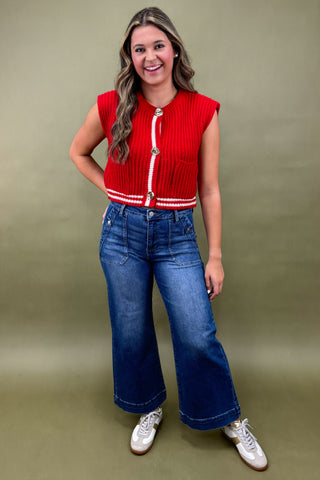 Woman wearing a red sleeveless top with white trim and blue jeans against a green background