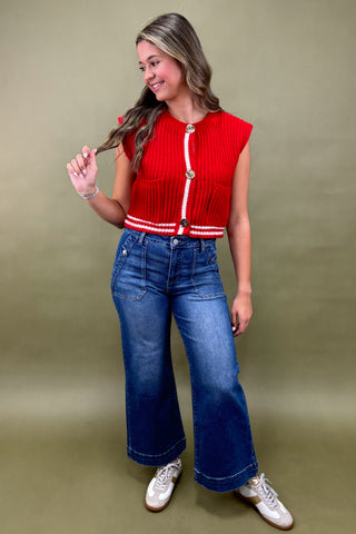 Woman wearing a red sleeveless top with white buttons and blue jeans against a green background