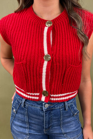 Red knitted vest with white stripe and gold buttons worn by a person against a green background