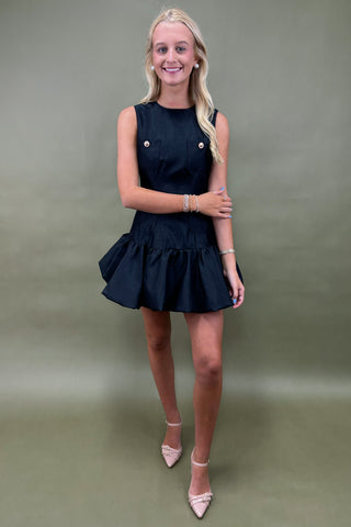 Woman wearing a black dress with gold buttons against a plain background