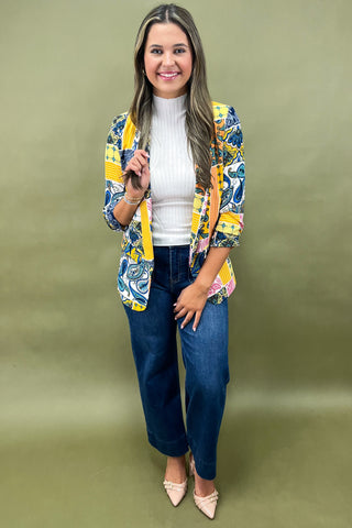 Woman wearing a colorful jacket, white shirt, blue jeans, and beige shoes on a green background