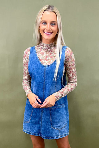 Woman wearing a blue denim dress over a floral top against a green background