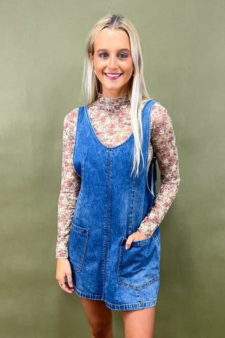 Woman wearing a blue denim pinafore dress over a floral top against a green background