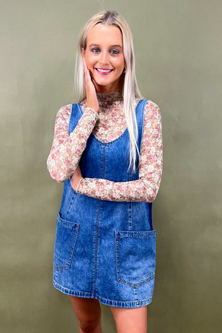 Woman wearing a blue denim dress over a floral long-sleeve shirt against a green background