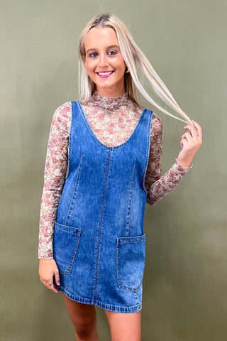 Woman wearing a denim dress over a floral long-sleeve shirt against a green background