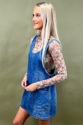 Woman wearing a denim pinafore dress with a floral shirt underneath on a green background