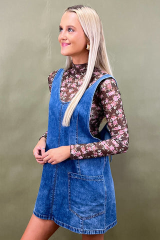 Woman wearing a blue denim dress over a floral blouse against a green background