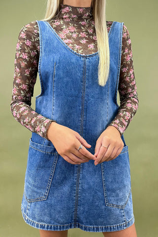 Person wearing a blue denim dress over a floral long-sleeve shirt on a green background