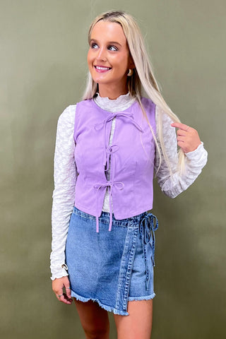 Woman wearing a purple top with a white lace overlay and denim skirt against a green background