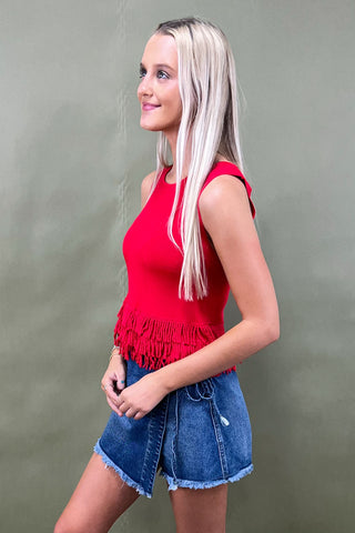 Woman wearing a red fringe top and denim shorts against a plain background