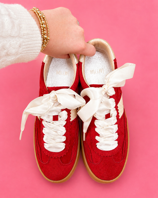 Red sneakers with white laces held by a hand against a pink background