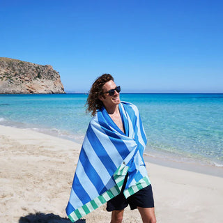 Person wrapped in a blue and green striped towel on a beach with clear blue water and sky.