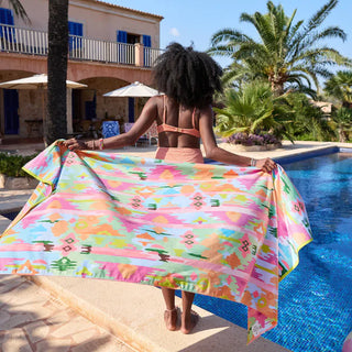 Woman holding a colorful towel by a poolside with a house and palm trees in the background
