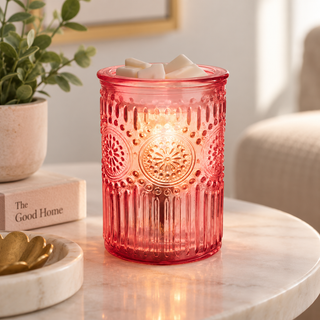 A pink textured glass candle holder with a lit candle inside, styled on a marble table with a potted plant and a book in the background.
