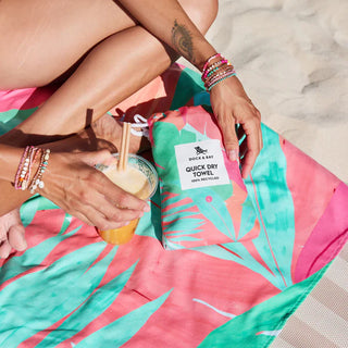 Person sitting on a colorful towel with a quick dry towel and drink, on a beach.