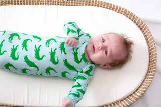 Baby lying on a white cushion in a wicker crib wearing a green and white outfit with alligator pattern.