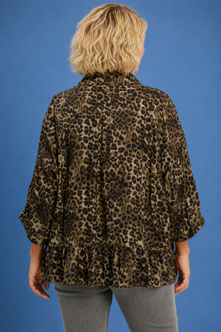 A woman viewed from the back wearing a leopard-print blouse with three-quarter sleeves and a ruffled hem, paired with gray pants, against a solid blue studio background.