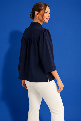 A woman wearing a navy blue blouse with three-quarter sleeves and a ruffled high neck, paired with white pants, standing against a solid blue studio background.