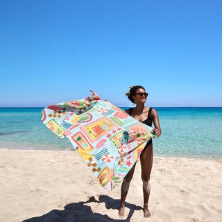 Woman holding a colorful towel on a beach with clear blue water and sky.