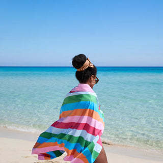 Person wrapped in a colorful towel on a beach with clear blue water and sky.