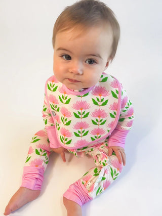 A baby wearing a pink and green floral print one-piece outfit with a matching bib, sitting against a plain white background.