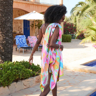 Woman in a colorful dress standing by a poolside with palm trees and lounge chairs in the background.