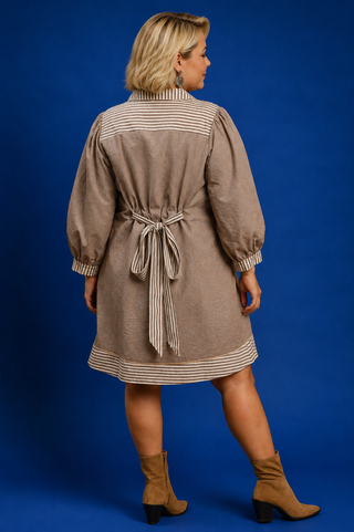 A woman shown from the back wearing a taupe-brown linen dress with beige and white striped details on the collar, sleeves, cuffs, and hem, tied at the waist with a matching tie belt. She is wearing tan ankle boots and standing against a solid blue studio background.