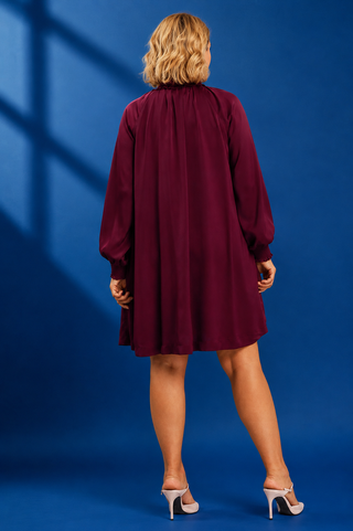 A woman viewed from behind wearing a long-sleeved, knee-length burgundy dress against a blue studio backdrop.