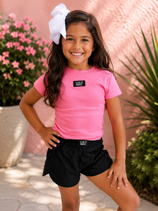 a bright pink short-sleeve t-shirt with a black rectangular 'LULU MAC' logo on the chest, paired with black athletic shorts