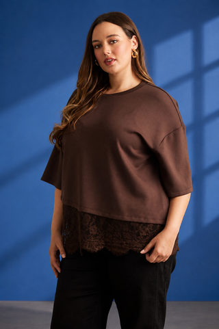 A woman modeling a brown short-sleeve top with a sheer black lace hem, paired with black pants, set against a blue studio backdrop with window shadow details.