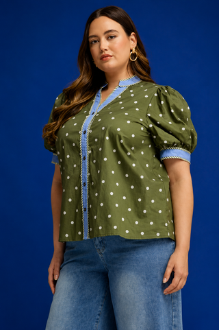 A woman modeling a short-sleeve button-front blouse with puff sleeves, olive green fabric, white polka dots, and light blue trim, paired with blue jeans against a solid blue studio background.