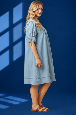 A woman modeling a light blue denim dress with puff sleeves and tie details at the cuffs, standing against a dark blue studio backdrop with window shadow overlays.