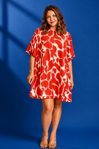 A woman modeling a short-sleeve, knee-length shift dress with an abstract red and off-white pattern, standing against a solid blue studio backdrop.