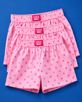 Three pairs of pink shorts with a darker pink polka dot pattern, stacked neatly on a solid blue background. Each pair features an elastic waistband with a pink label that reads 'LULU MAC'.