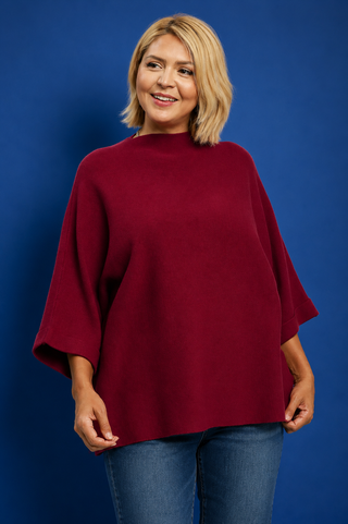 A woman with blonde hair wearing a loose-fitting, deep burgundy knit poncho top and blue jeans, standing against a solid blue studio background.