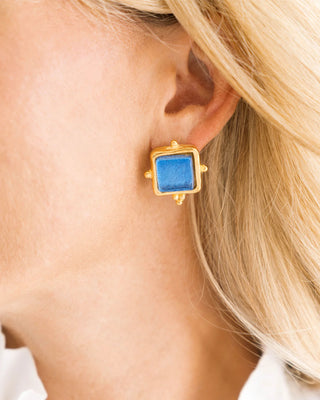 Gold earring with a blue gemstone worn by a person with blonde hair.