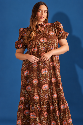 A woman modeling a brown bohemian-style midi dress with puff sleeves, a high ruffled collar, and an all-over floral print in pink, orange, and blue