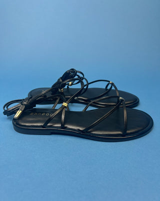 A pair of black leather flat sandals with thin strappy details and small gold hardware, against a solid blue background.