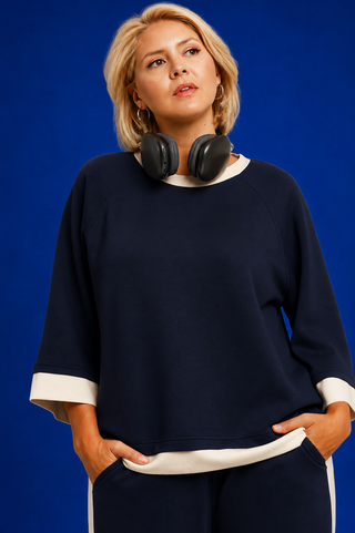 A woman wearing a navy blue sweatshirt with contrasting white ribbed trim at the collar, cuffs, and hem, paired with matching navy pants. She has black over-ear headphones around her neck and is posed against a solid blue studio background.