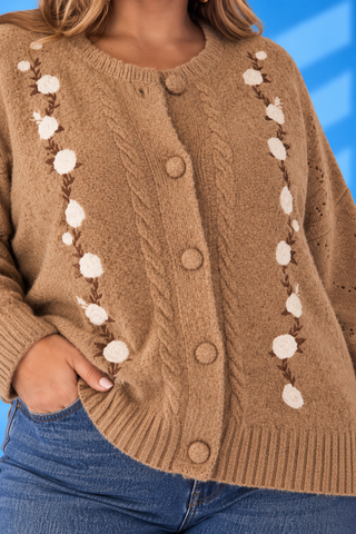 A close-up of a person wearing a tan cable-knit cardigan sweater with vertical floral embroidery and blue jeans