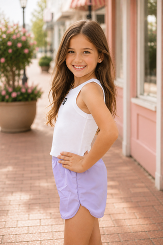 A young girl stands outdoors on a sunlit walkway wearing a light lavender sleeveless tank top and matching shorts