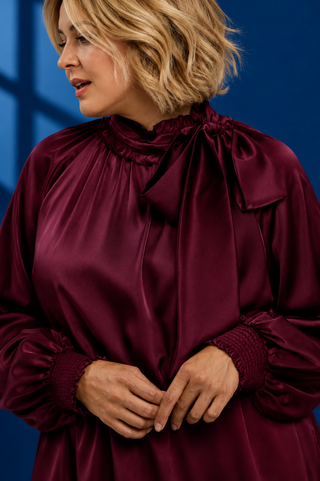 A woman wearing a long-sleeved satin blouse in deep burgundy with a high ruffled collar and a large bow at the neck, set against a dark blue background.