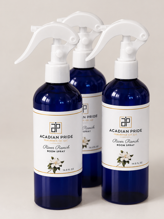 Three cobalt blue spray bottles with white trigger tops and white labels reading Acadian Pride River Ranch Room Spray, arranged on a light beige background.
