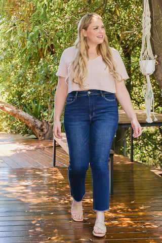 Woman wearing blue jeans and a light-colored top on a wooden deck with greenery in the background