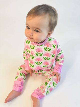 A baby sitting on a white studio background wearing a pink and green floral print long-sleeve romper and matching pants.