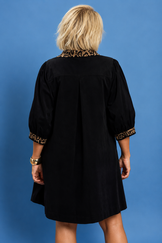 A woman viewed from the back wearing a black short-sleeve dress with leopard-print trim at the collar and cuffs, standing against a solid blue background.